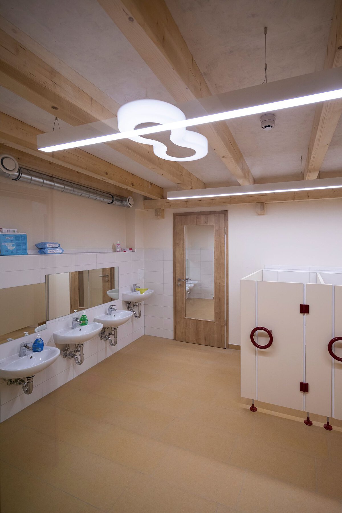 Waschraum im Kindergarten Am Wald- Klinikum mit drei Waschbecken und zwei Kabinen Waschraum. An der linken Wand hängen drei kleine Waschbecken mit Spiegeln darüber. Auf den Waschbecken stehen bunte Seifenspender. Rechts sind zwei helle Kabinen mit roten Griffen. In der Mitte steht eine Holztür. Die Decke hat sichtbare Holzbalken und eine Lampe in einer besonderen Form. Der Boden ist hellbraun