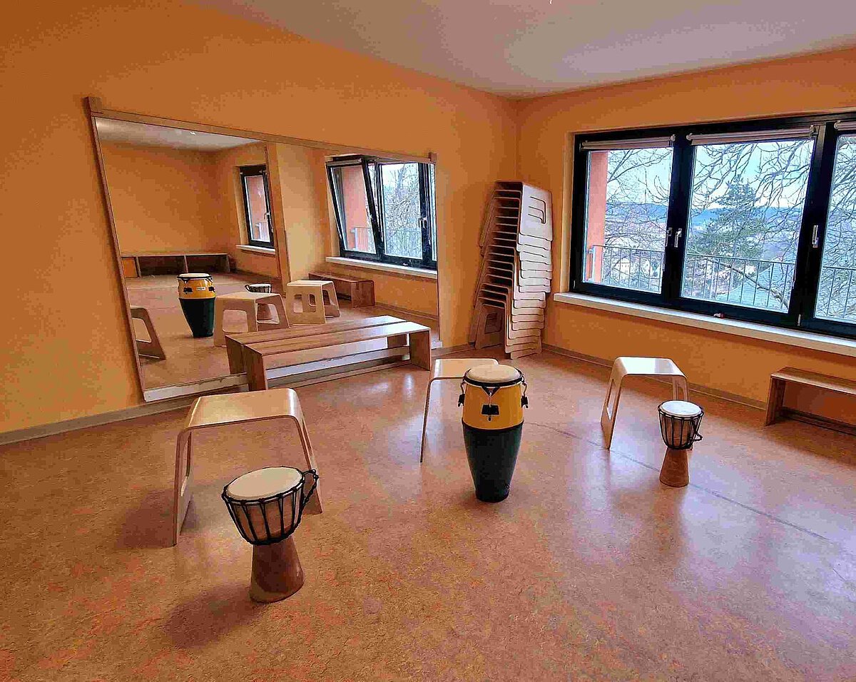 Rhythmikraum im Kindergarten mit großen Fenstern, Spiegelwand und mehreren Trommeln auf dem Boden. Im Raum stehen kleine Holzstühle und ein Podest, daneben ein Stapel weiterer Stühle. Der Raum ist hell und freundlich gestaltet mit einem großen Instrumentarium für Kinder.
