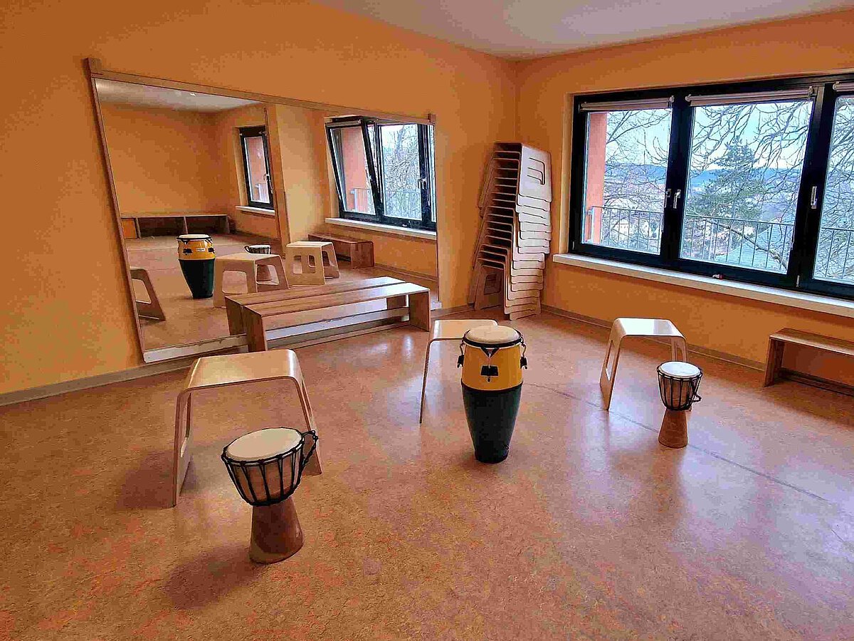 Rhythmikraum im Kindergarten Burgweg – Musik und Bewegung für Kinder Rhythmikraum im Kindergarten mit großen Fenstern, Spiegelwand und mehreren Trommeln auf dem Boden. Im Raum stehen kleine Holzstühle und ein Podest, daneben ein Stapel weiterer Stühle. Der Raum ist hell und freundlich gestaltet mit einem großen Instrumentarium für Kinder.