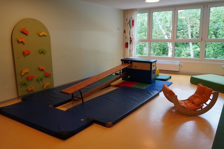 Bewegungsraum mit Turnmatten, Kletterwand, Balancierbank und Spielmodulen für Kinder, großer Fensterfront.