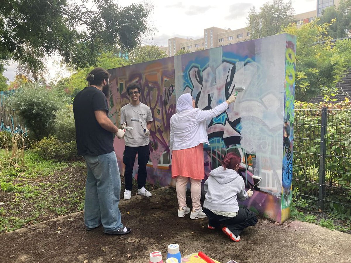 Mehrere Personen arbeiten gemeinsam an einer bunt bemalten Graffiti-Wand im Freien. Sie tragen Handschuhe und nutzen Farbrollen, um die Wand zu gestalten. Im Hintergrund sind Bäume und ein Wohngebäude zu sehen.