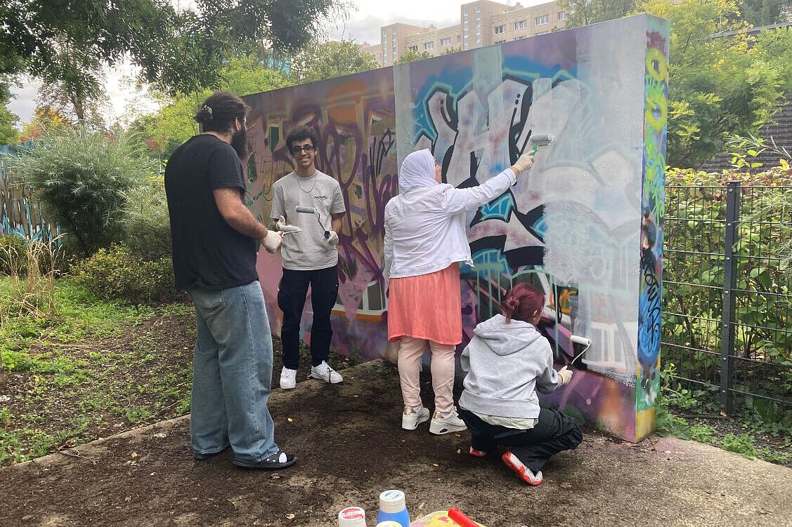 Mehrere Personen arbeiten gemeinsam an einer bunt bemalten Graffiti-Wand im Freien. Sie tragen Handschuhe und nutzen Farbrollen, um die Wand zu gestalten. Im Hintergrund sind Bäume und ein Wohngebäude zu sehen.
