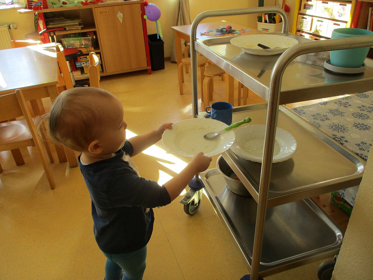 Selbstständigkeit - Tisch abräumen - Kindergarten Windischholzhausen Ein jüngeres Kind bringt selbstständig den leeren Teller vom Mittagessen auf den Essenwagen.
