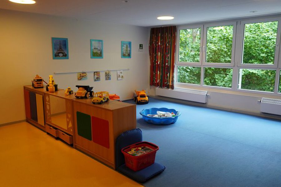 Bauraum in der Krippe mit niedrigen Regalen, Spielzeugfahrzeugen, Bauelementen und weicher Spielfläche vor großer Fensterfront.