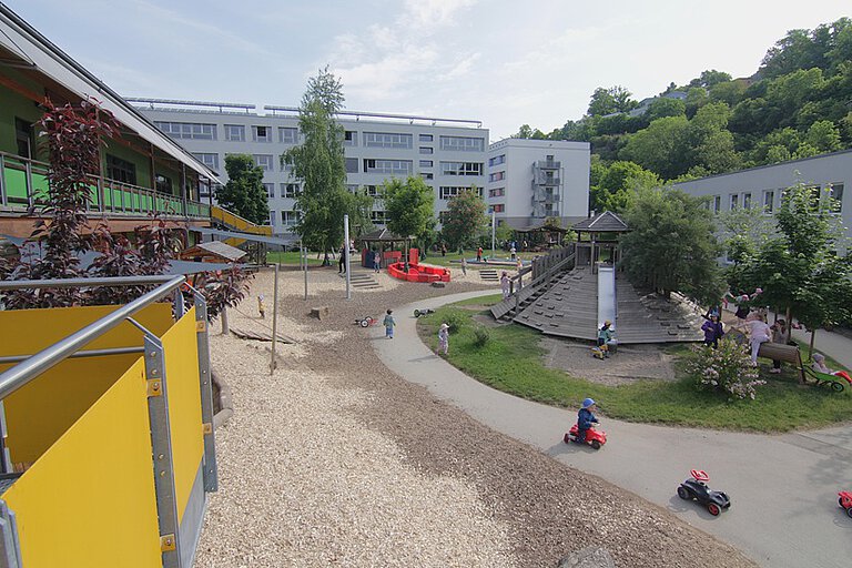ein Blick auf den großen Garten der Kita Sportforum im Garten des Kindergartens steht ein holzverkleideter Berg mit einer Rutsche, Kinder fahren mit Fahrzeugen auf einer asphaltierten Straße, die darum führt