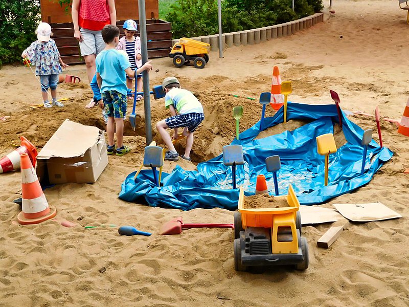 Kinder spielen im Sandkasten mit Schaufeln und haben ein tiefes Loch gegraben. Viele Sandspielzeuge und Fahrzeuge umgeben sie.