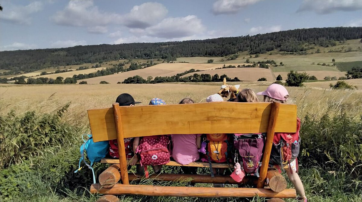 Kinder sitzen mit Rucksäcken auf einer Holzbank und blicken auf Felder und Wald