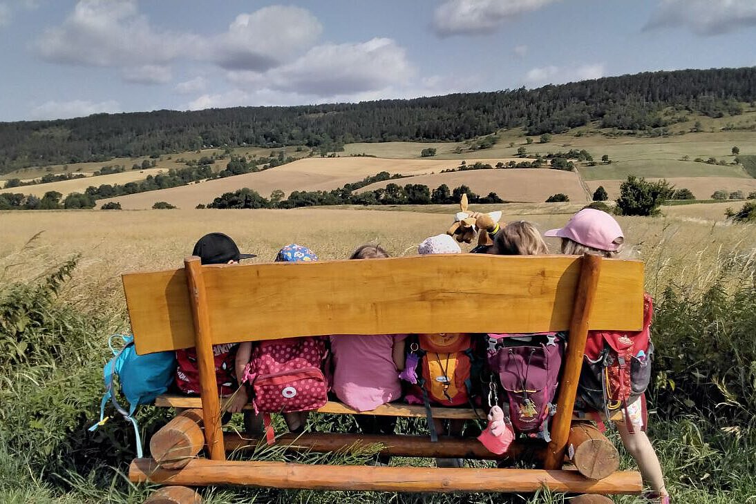 Kinder sitzen mit Rucksäcken auf einer Holzbank und blicken auf Felder und Wald