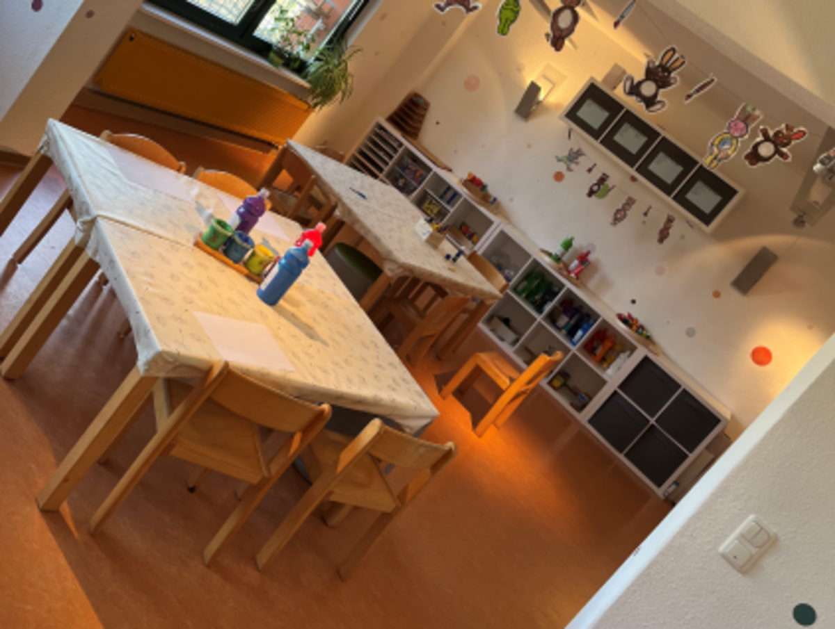 Heller Kreativraum im Kindergarten Burgweg mit großen Tischen und Holzstühlen, Bastelmaterialien wie Stifte und Wasserfarben auf den Tischen, Regalen mit bunten Bastelutensilien und dekorativen Figuren an der Decke.