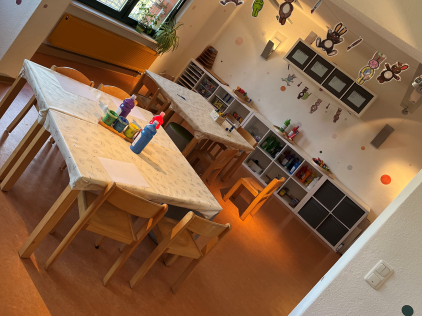 Kreativraum im Kindergarten Burgweg – Basteln und Malen in kindgerechter Umgebung Heller Kreativraum im Kindergarten Burgweg mit großen Tischen und Holzstühlen, Bastelmaterialien wie Stifte und Wasserfarben auf den Tischen, Regalen mit bunten Bastelutensilien und dekorativen Figuren an der Decke.