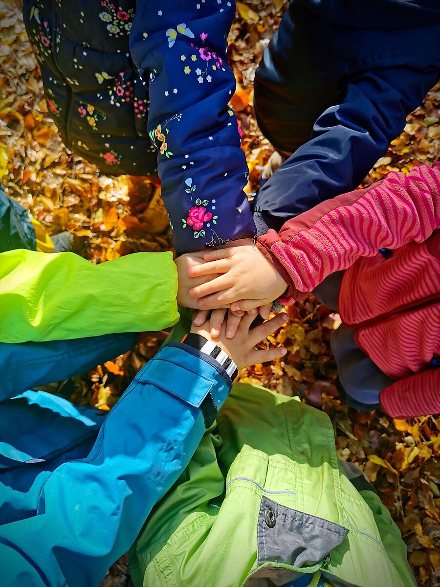 Mehrere Kinder legen ihre Hände im Kindergarten Burgweg übereinander auf einem Boden voller Herbstlaub. Die Kinder tragen bunte Jacken in Blau, Grün und Pink, was ein Gefühl von Teamgeist und Gemeinschaft vermittelt.