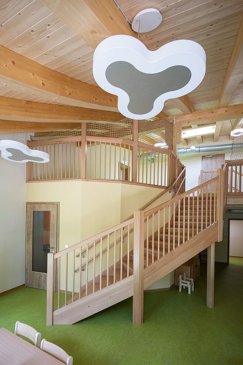 Innenraum mit einer Holztreppe, die nach oben führt. Die Treppe hat ein Geländer aus Holz. An der Decke sind große Holzbalken und Lampen in Form von Wolken. Der Boden ist grün. Im Hintergrund sieht man eine Tür und einen Bereich mit hellen Wänden. Der Raum wirkt offen und freundlich