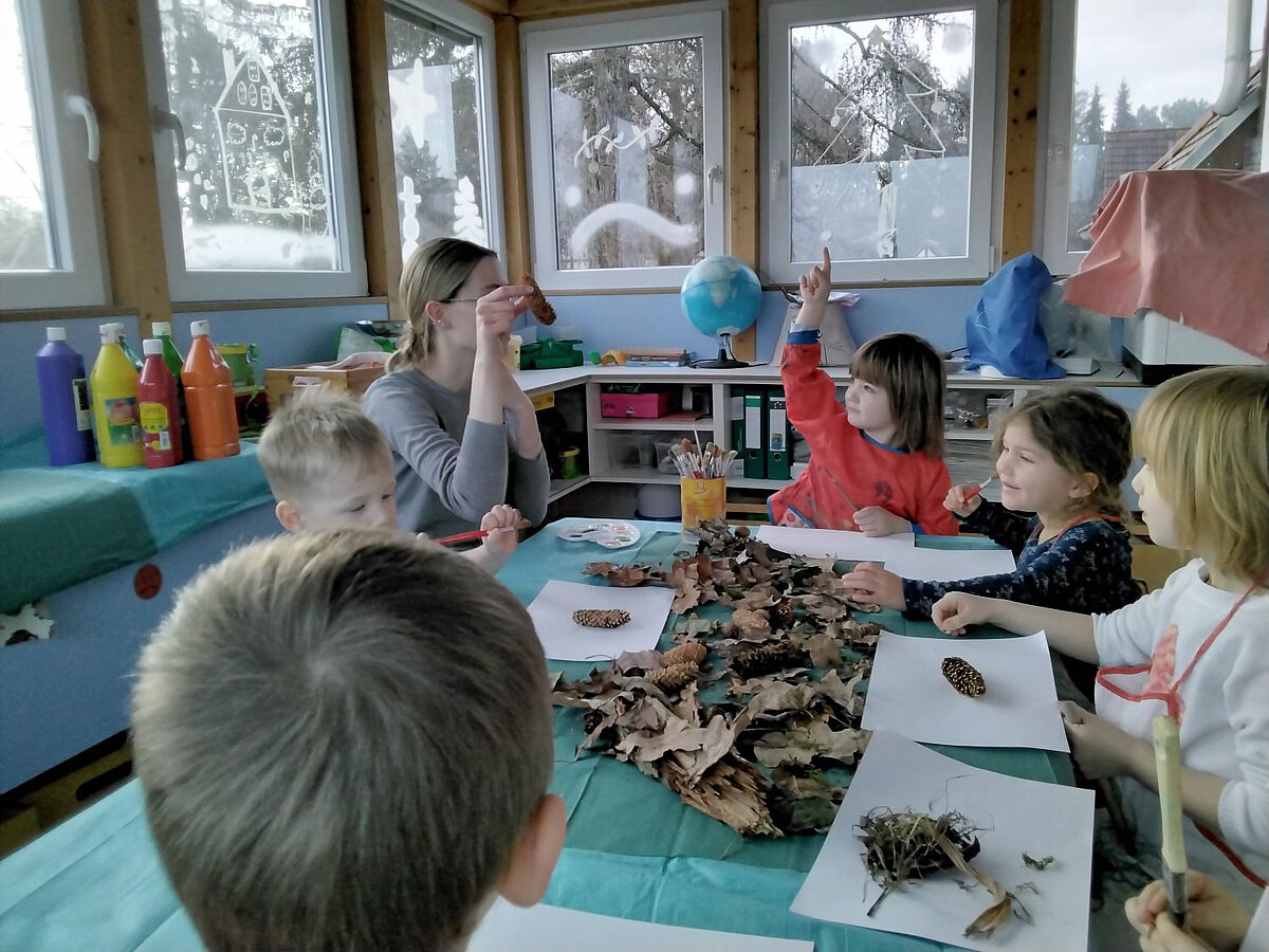 Naturmaterialien - Kreativität - Forscherraum - Kindergarten Windischholzhausen Eine Erzieherin sitzt mit Kindern an einem Tisch und hält einen Tannenzapfen nach oben. Der Tisch ist voller Naturmaterialien. Ein Kind meldet sich, um der Erzieherin zu sagen, dass sie einen Tannenzapfen in der Hand hält