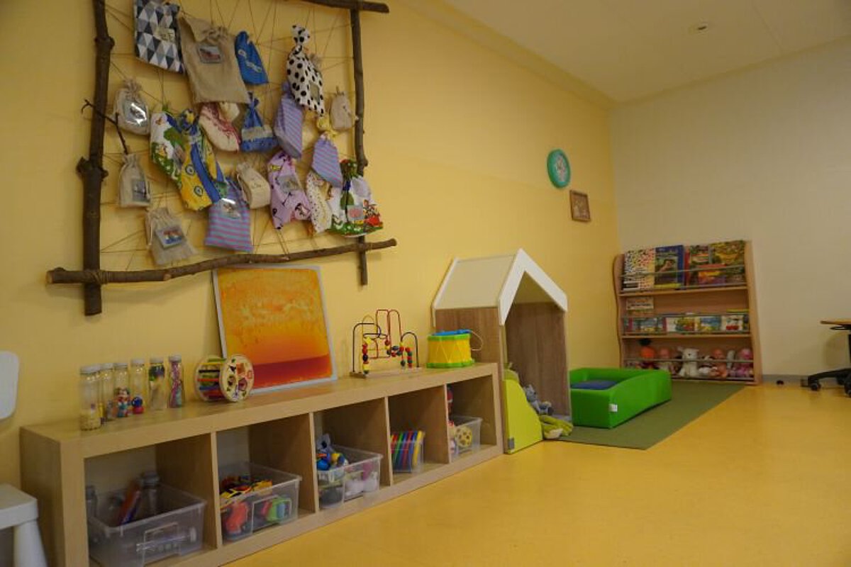 Spiel- und Kreativbereich in der Krippe mit Regalen voller Materialien, Wandgestaltung mit Bastelarbeiten, Spielhaus und Bücherregal.