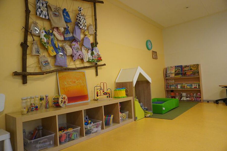 Spiel- und Kreativbereich in der Krippe mit Regalen voller Materialien, Wandgestaltung mit Bastelarbeiten, Spielhaus und Bücherregal.