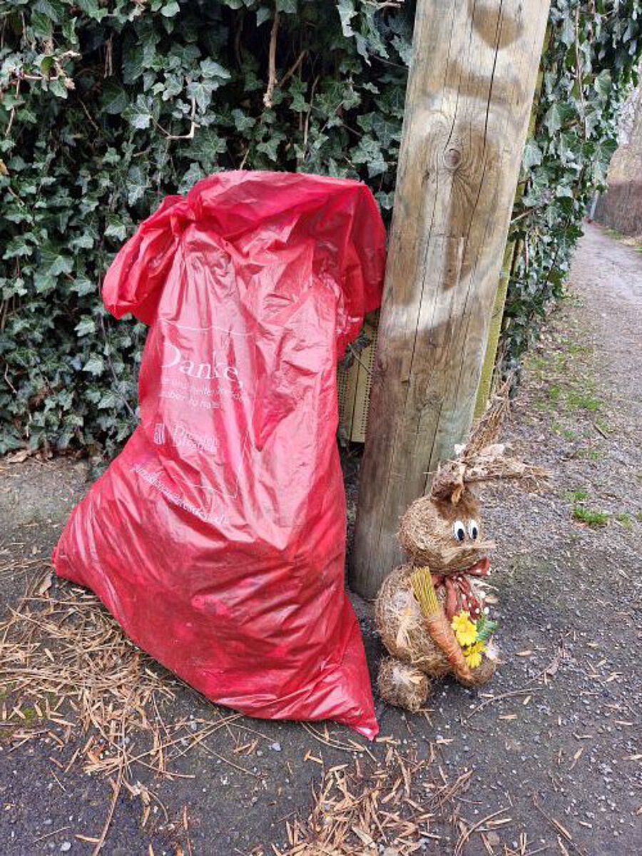 „Großer roter Müllsack lehnt an einem hölzernen Pfosten neben einem dekorativen Hasen aus Naturmaterialien, im Hintergrund ein mit Efeu bewachsener Zaun und ein befestigter Weg.“