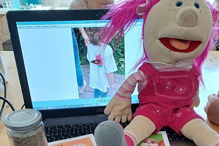 Maskottchen Kirschi der Kindertagesstätte Kirschallee sitzt auf dem Laptop mit Materialien