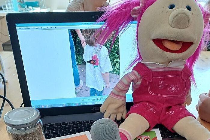 Maskottchen Kirschi der Kindertagesstätte Kirschallee sitzt auf dem Laptop mit Materialien