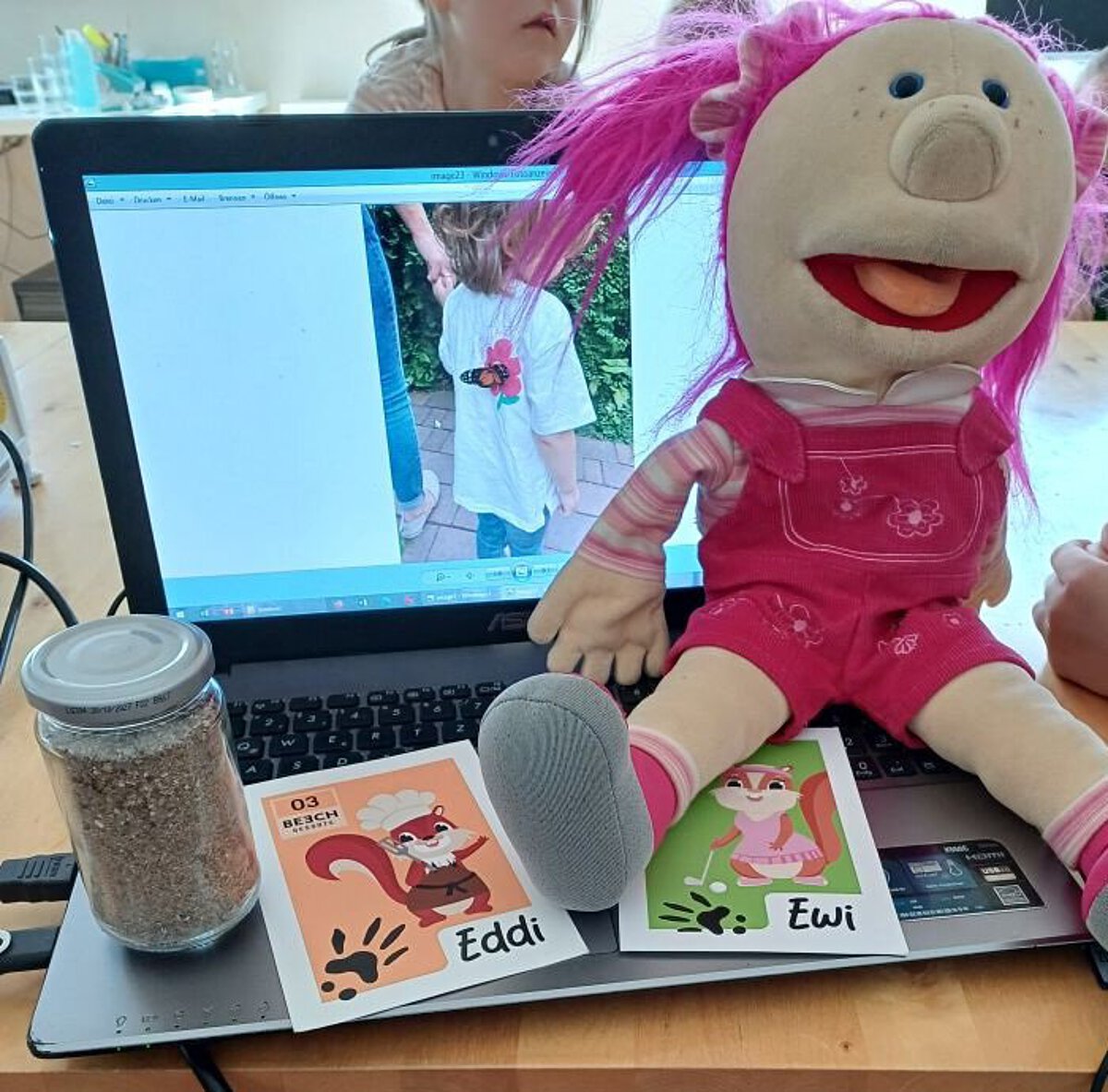 Maskottchen Kirschi der Kindertagesstätte Kirschallee sitzt auf dem Laptop mit Materialien