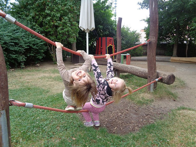 Fröhliche Kinder - Spaß beim Klettern - Lachen - Kindergarten Windischholzhausen Das Bild zeigt zwei Kinder, die auf einem Spielplatz zwischen zwei roten Seilen hängen und sich daran festhalten, während im Hintergrund Bäume, ein Sonnenschirm und ein rotes Spielhaus zu sehen sind