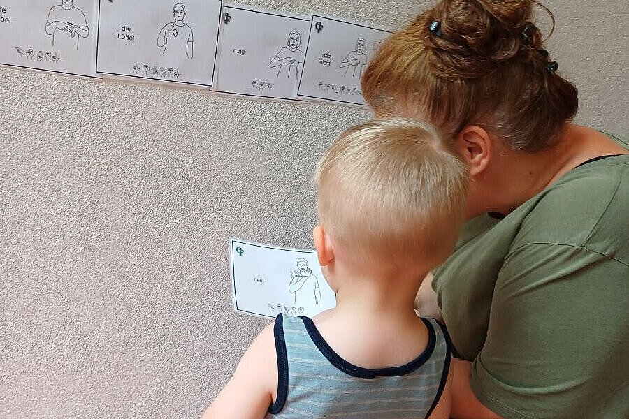 Gebärdenschatzkiste im Waldkindergarten Ottendorf-Okrilla Kind betrachtet gemeinsam mit einer pädagogischen Fachkraft Gebärden-Bildkarten an einer Wand im Waldkindergarten Ottendorf-Okrilla
