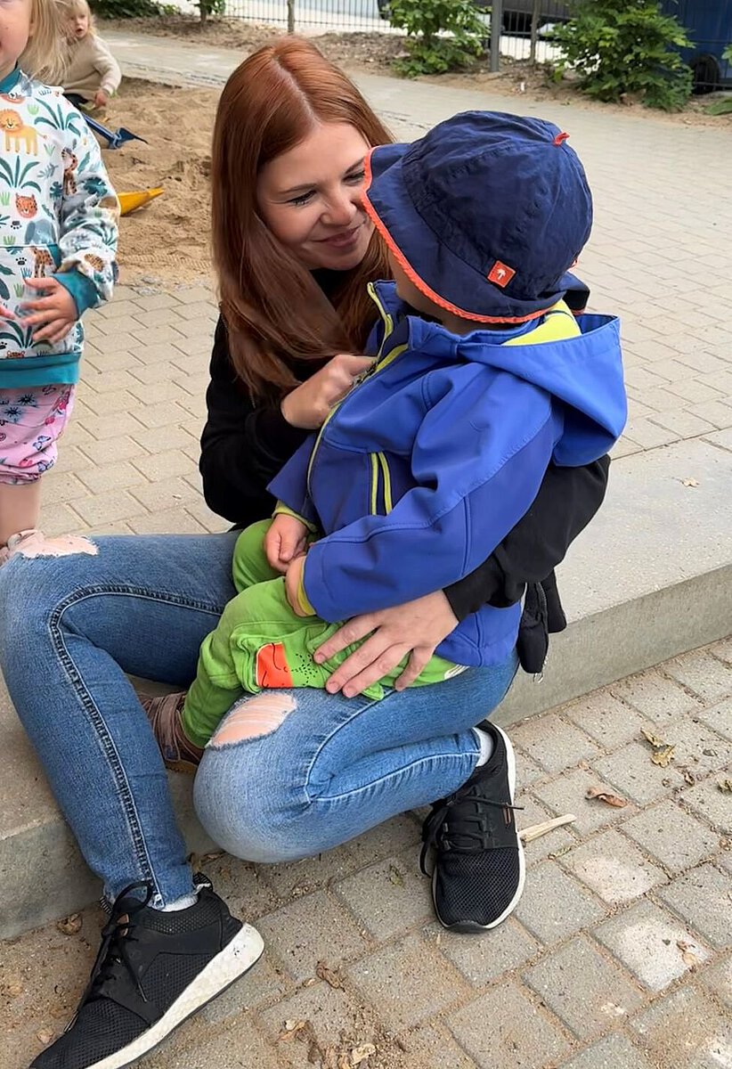 Eine pädagogische Fachkraft sitzt mit einem Kind im Außenbereich der Kita und widmet ihm ihre ungeteilte Aufmerksamkeit. Verlässliche Beziehungen geben Kindern Sicherheit und stärken ihre emotionale Entwicklung.