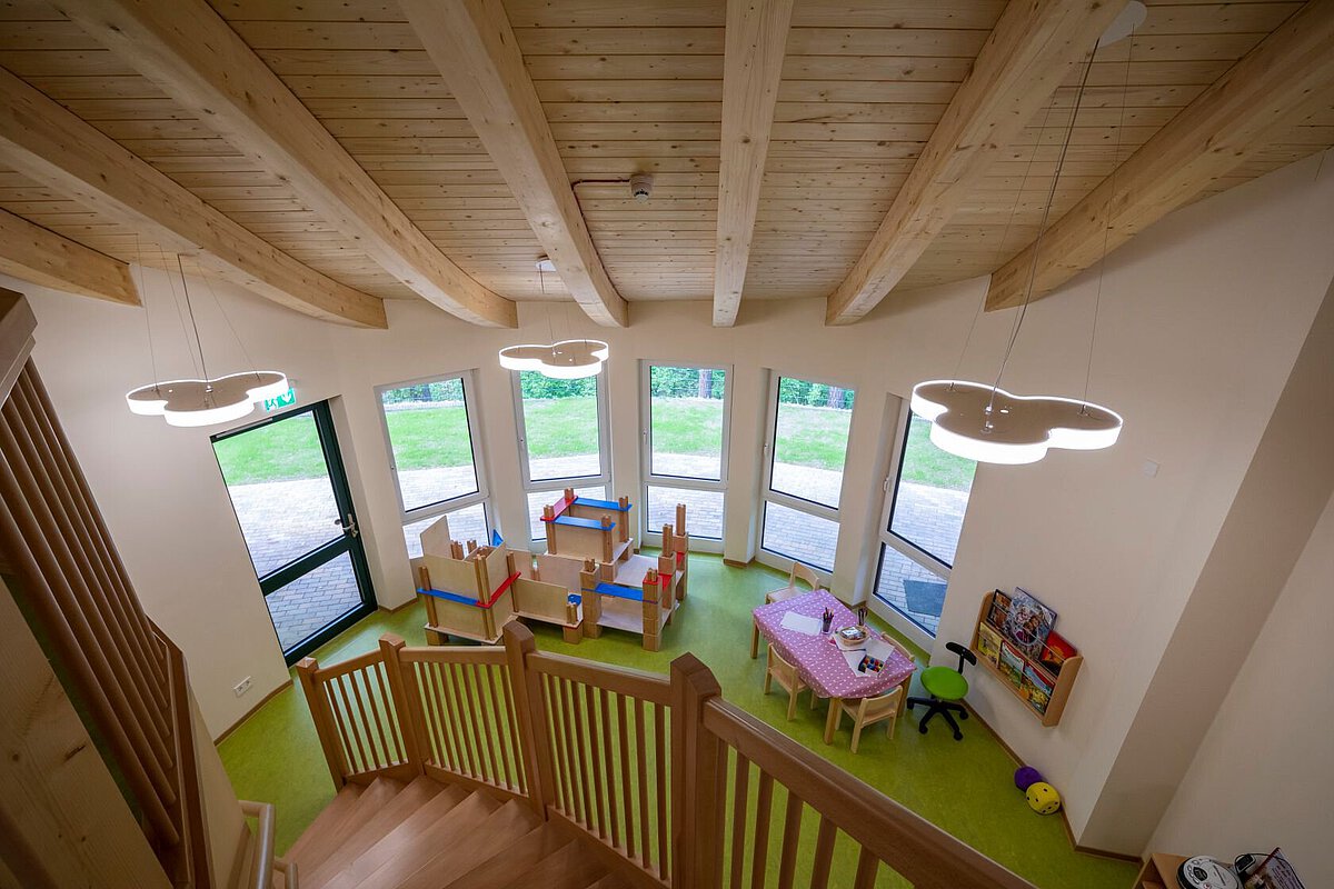 heller Raum mit einer Holzdecke und großen Fenstern . Der Boden ist grün. In der Mitte stehen kleine Holztische und Stühle. Auf einem Tisch liegt eine rot-weiß karierte Tischdecke. An der Wand rechts ist ein Regal mit Spielzeug. Von oben führt eine Holztreppe in den Raum. An der Decke hängen Lampen in Wolkenform. Draußen vor den Fenstern ist eine Wiese zu sehen