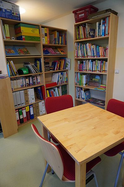 Bibliotheks‑ und Arbeitsraum mit Bücherregalen voller Kinderbücher, Ordnern sowie Tisch mit Stühlen.