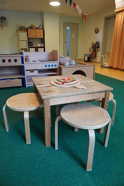 Rollenspielraum in der Krippe mit kleinem Tisch und Hockern, Spielküche und Materialien für gemeinsames Spielen.