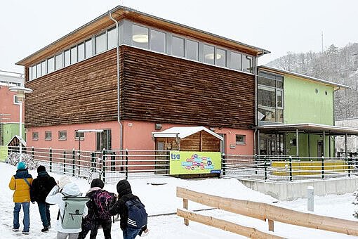 Winterlicher Ankunftsmoment an der Kita Sportforum Mehrere Kinder laufen bei verschneitem Winterwetter auf das Gebäude der Kita zu, das aus Holz- und Farbfassaden besteht.