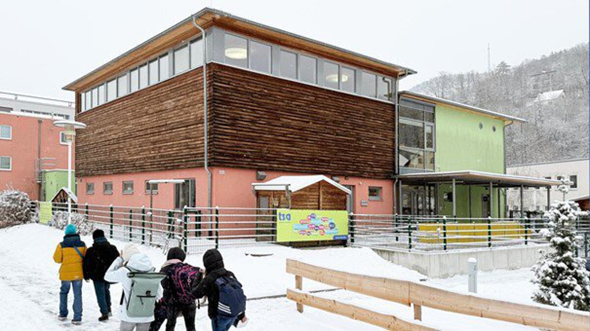 Mehrere Kinder laufen bei verschneitem Winterwetter auf das Gebäude der Kita zu, das aus Holz- und Farbfassaden besteht.