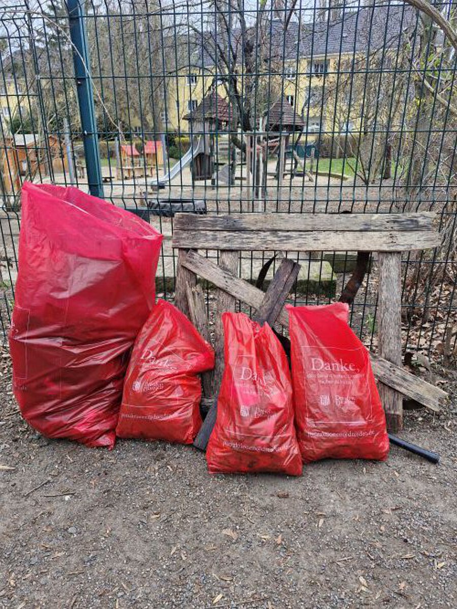 „Mehrere rote Müllsäcke stehen gefüllt vor einem Zaun neben alten Holzbrettern, im Hintergrund ein eingezäunter Spielplatz und Wohngebäude.“