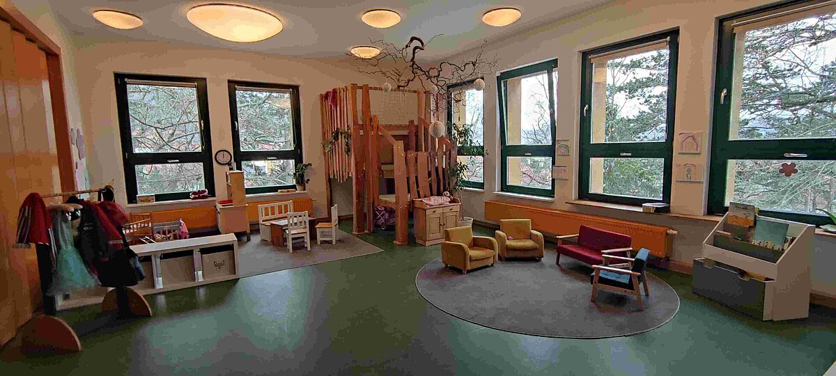 Rollenspielzimmer im Kindergarten Burgweg mit großen Fenstern, einer Hochebene aus Holz, einer Spielecke mit Holzspielmöbeln, kleinen Sesseln und Sofas auf einem Teppich sowie einer Garderobe mit Kostümen. Der Raum ist hell und freundlich gestaltet.