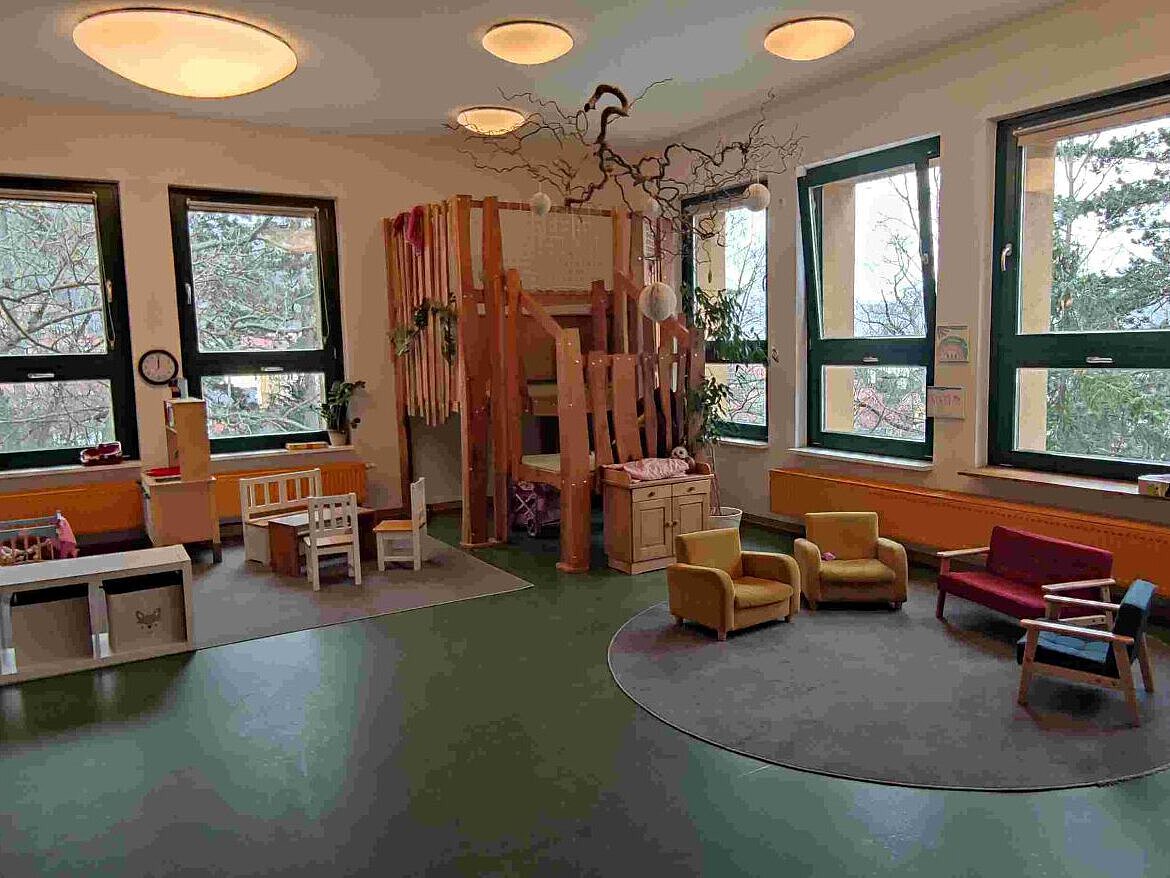 Rollenspielzimmer im Kindergarten Burgweg – Fantasievolles Spielen in gemütlicher Umgebung Rollenspielzimmer im Kindergarten Burgweg mit großen Fenstern, einer Hochebene aus Holz, einer Spielecke mit Holzspielmöbeln, kleinen Sesseln und Sofas auf einem Teppich sowie einer Garderobe mit Kostümen. Der Raum ist hell und freundlich gestaltet.