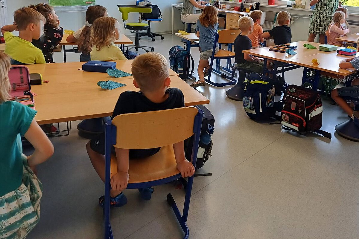 Kinder sitzen in einem Klassenzimmer an Gruppentischen und hören einer Lehrkraft zu.