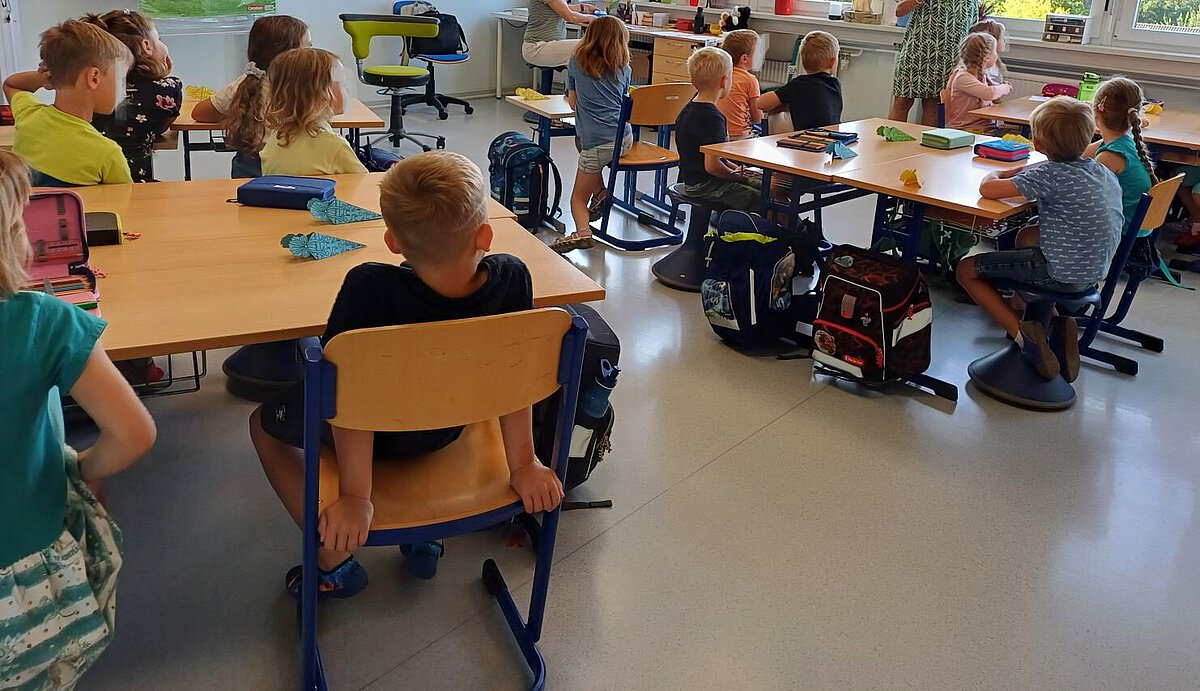 Kinder sitzen in einem Klassenzimmer an Gruppentischen und hören einer Lehrkraft zu.