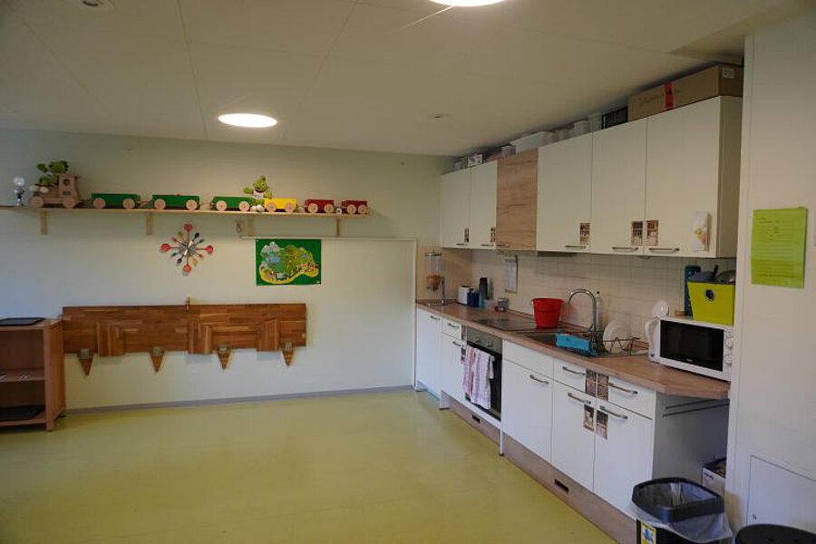 Essbereich im Kindergarten mit Kinderküche, Arbeitsflächen, Schränken und Wandregalen in einem hellen Raum.