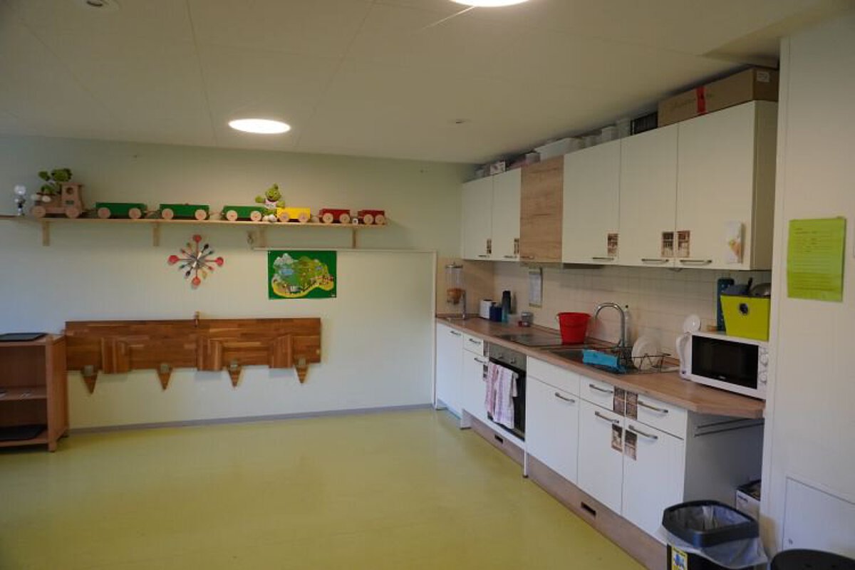 Essbereich im Kindergarten mit Kinderküche, Arbeitsflächen, Schränken und Wandregalen in einem hellen Raum.