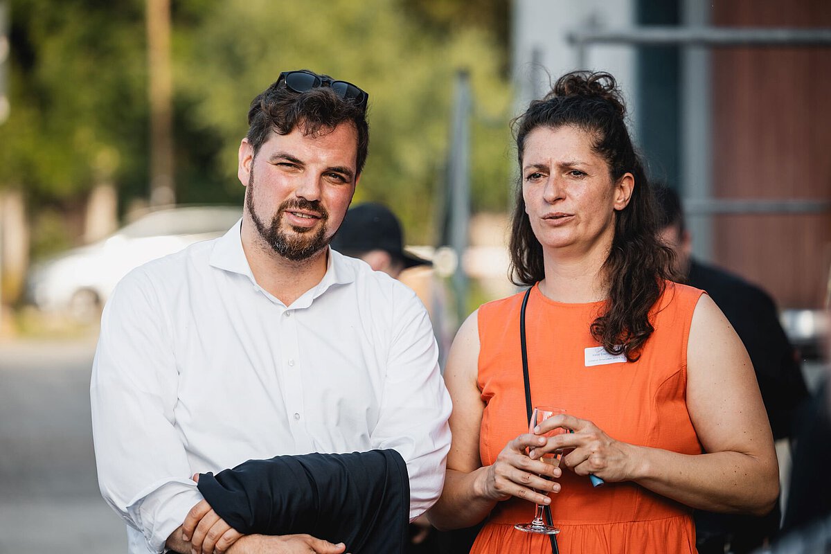Christian Schleußner und Katja Eisenschmidt stehen im Außenbereich einer Veranstaltung, unterhalten sich und halten Getränke in der Hand.