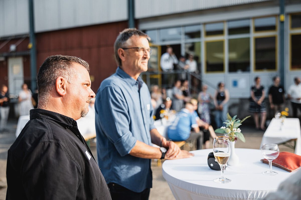 Personen stehen an Stehtischen vor einem Gebäude und verfolgen eine Veranstaltung im Außenbereich.
