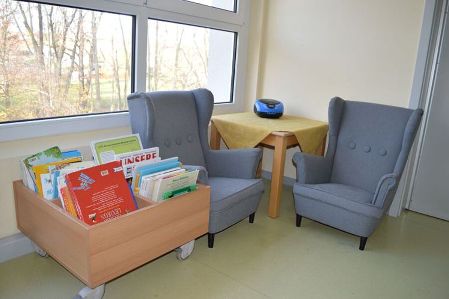 Leseecke mit zwei Polstersesseln, Bücherwagen mit Kinderbüchern und kleinem Tisch am Fenster.