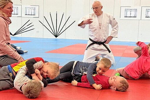 Kinder üben gemeinsam spielerische Judo-Bewegungen auf einer roten und blauen Matte, angeleitet vom Trainer im Judogi.