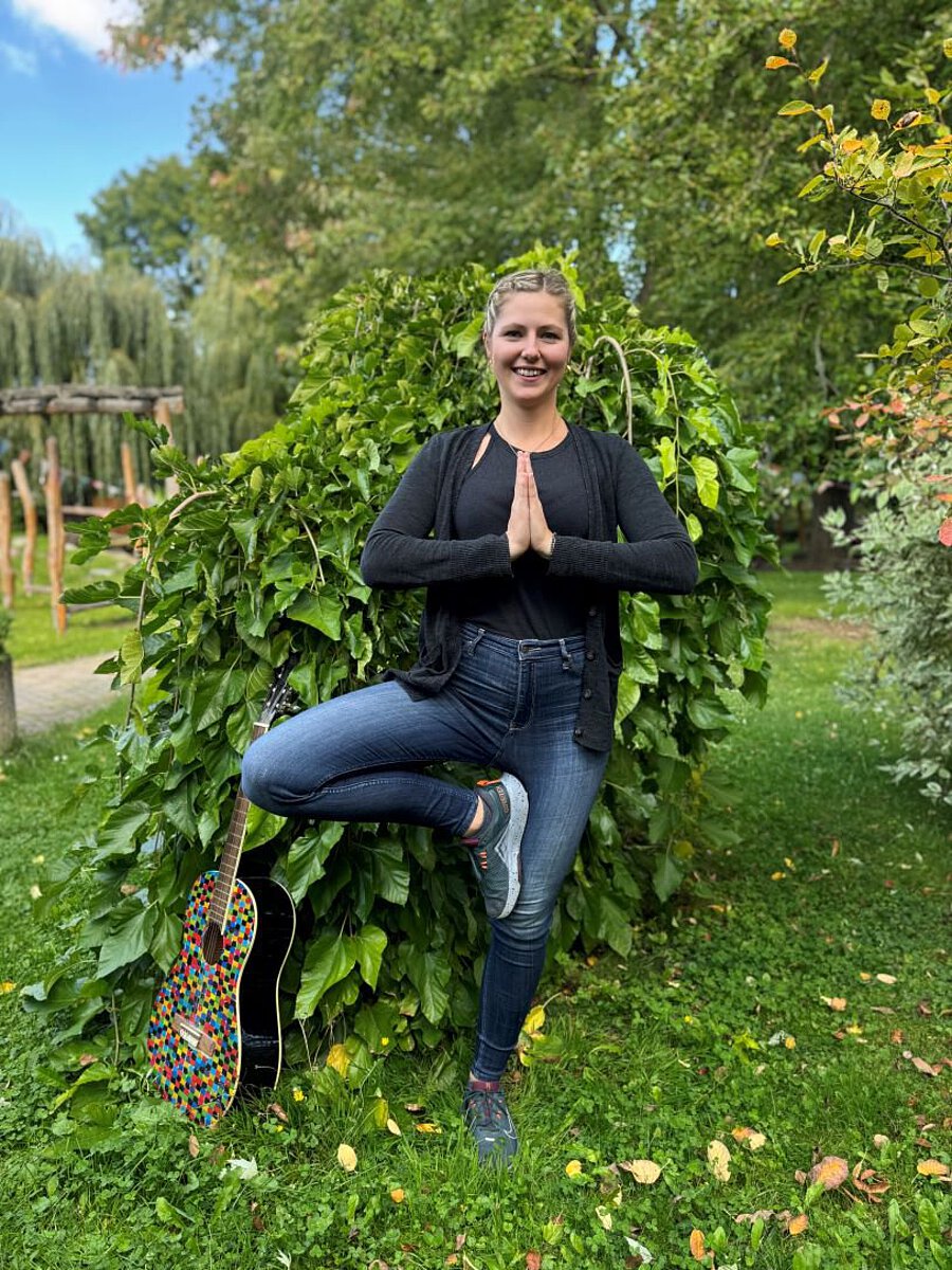 Erzieherin Laura Stegmann steht im Freien vor einem grünen Busch, zeigt eine Yoga-Pose und trägt eine schwarze Jacke mit Jeans. Neben ihr lehnt eine bunt bemalte Gitarre – ein Symbol für kreative pädagogische Arbeit.
