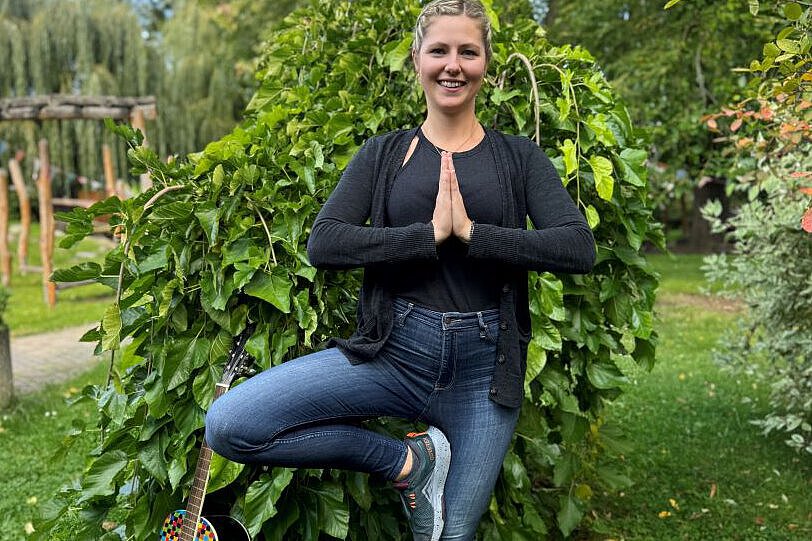 Erzieherin Laura Stegmann steht im Freien vor einem grünen Busch, zeigt eine Yoga-Pose und trägt eine schwarze Jacke mit Jeans. Neben ihr lehnt eine bunt bemalte Gitarre – ein Symbol für kreative pädagogische Arbeit.
