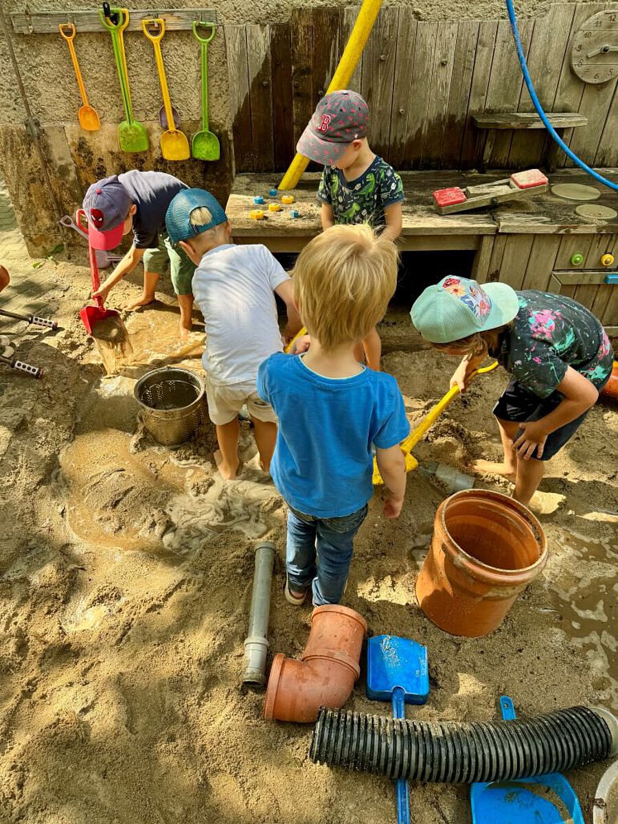 Kinder spielen gemeinsam an einer Wasser- und Matschanlage im Außengelände der Kita. Beim Bauen, Schaufeln und Experimentieren sammeln sie Erfahrungen, stärken ihr Selbstvertrauen und lernen miteinander zu kooperieren.