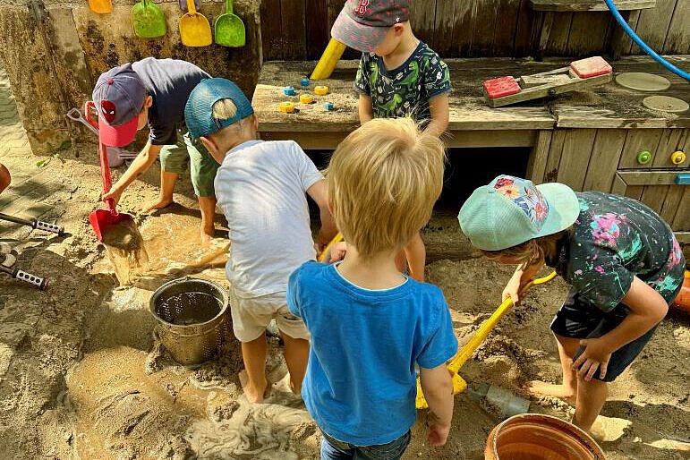 Kinder spielen gemeinsam an einer Wasser- und Matschanlage im Außengelände der Kita. Beim Bauen, Schaufeln und Experimentieren sammeln sie Erfahrungen, stärken ihr Selbstvertrauen und lernen miteinander zu kooperieren.