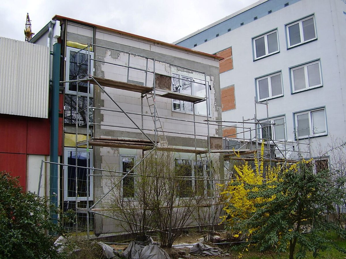 Umbau des Verbindungsbaus am Akademiehotel mit Gerüst, freigelegter Fassade und Baustelle im Außenbereich mit Sträuchern und gelb blühender Pflanze