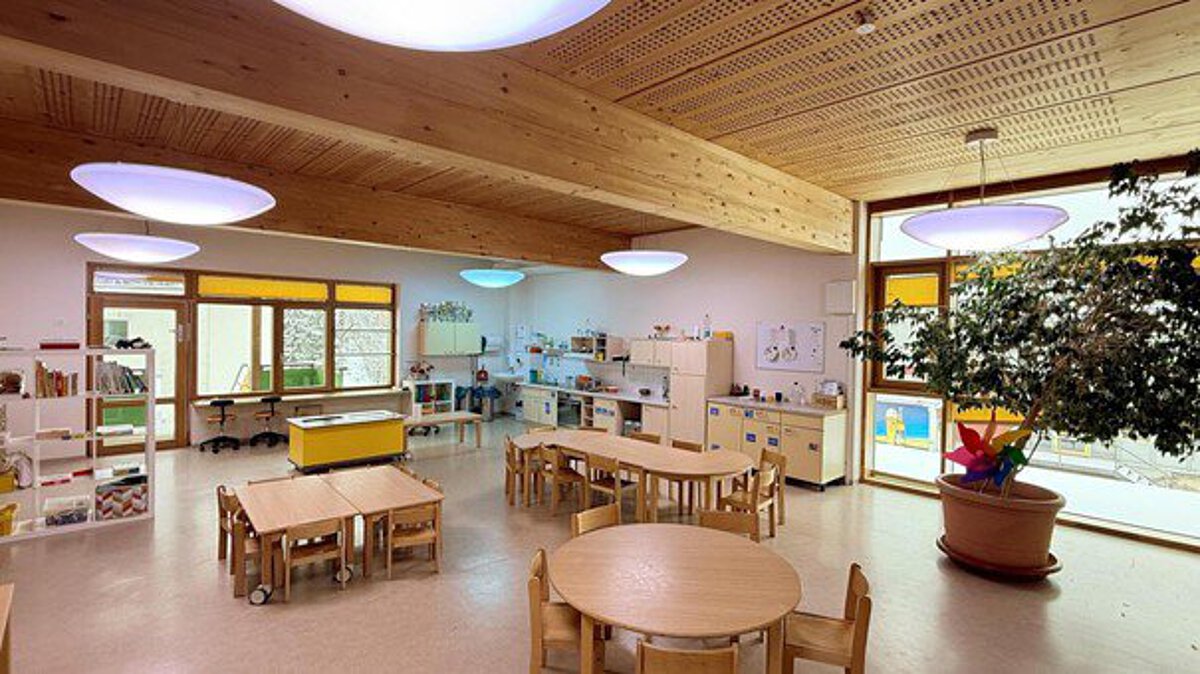 Großer, freundlicher Kita-Raum mit Holzmöbeln, Bastel- und Spielebereichen sowie großen Fenstern mit Blick nach draußen.