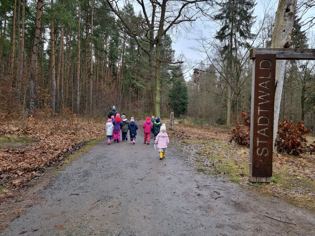 Das Bild zeigt einen Waldweg. Auf dem Weg gehen mehrere Kinder in bunten Jacken und Mützen. Vorne rechts steht ein Holzschild mit der Aufschrift „STADTWALD“. Links und rechts vom Weg sind viele Bäume und braune Blätter auf dem Boden. Der Himmel ist grau