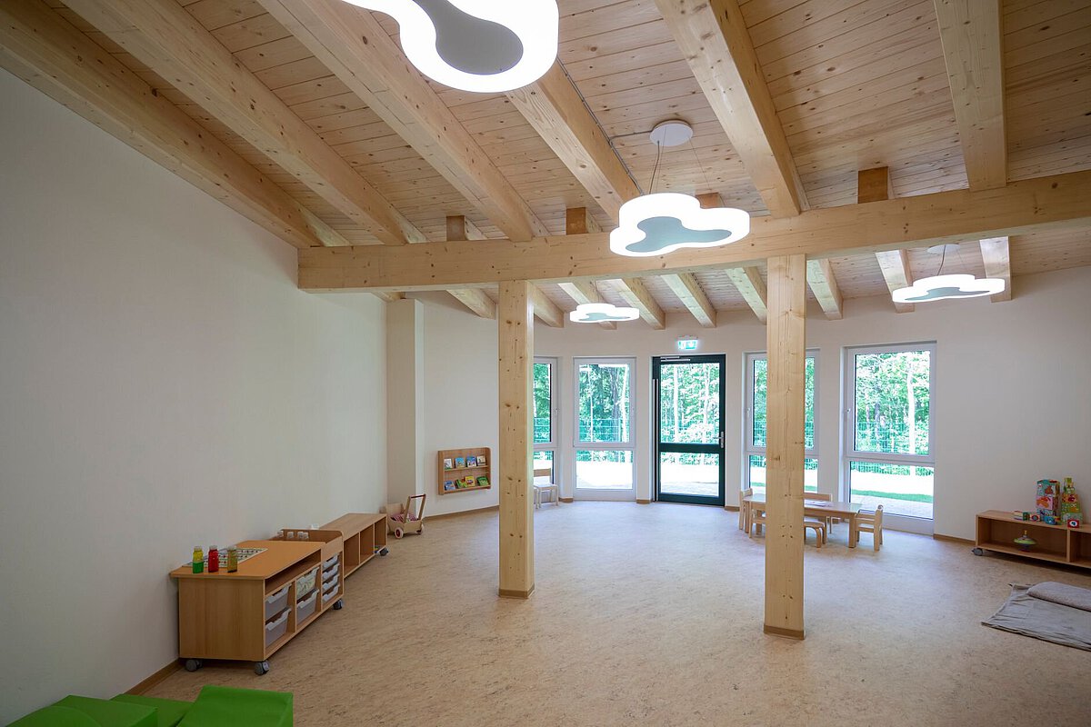 heller Raum mit Holzdecke und großen Fenstern. An der Decke hängen mehrere Lampen in Form von Wolken. Im Raum stehen kleine Holzmöbel, wie Regale und Tische. Auf den Regalen liegen Spielmaterialien. Der Boden ist hell und der Raum wirkt freundlich und offen